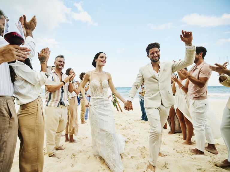 Newlyweds walk hand in hand down a beach while their loved ones cheer and celebrate. 
