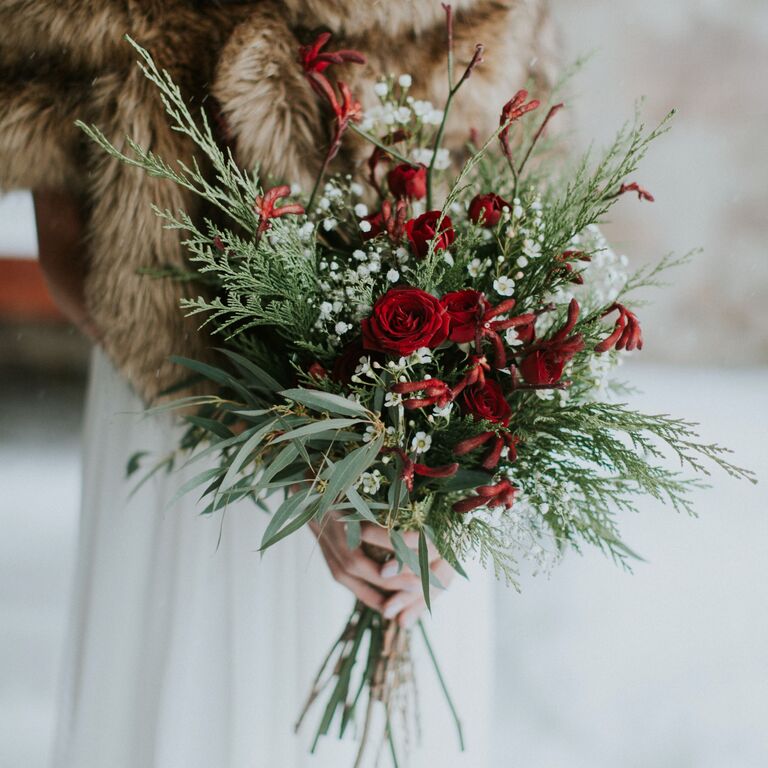 bride wearing brown fur wrap holds winter wedding bouquet with red spray roses and evergreen branches