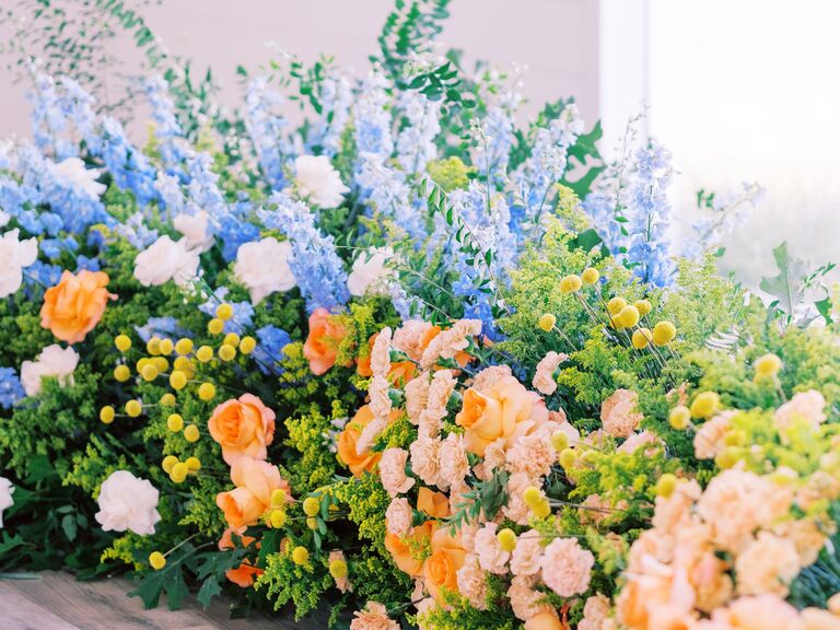 oversized wedding flower arrangement with blue delphinium, yellow craspedia, orange carnations and roses and greenery